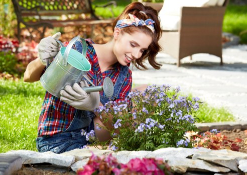 A young woman with brown hair tied back with a colorful headband, wearing a plaid shirt and grey gardening gloves, is kneeling on a well-maintained garden lawn near a flower bed. She is using a small metal hand trowel to tend to a bush of light purple flowers. Behind her, there is a paved patio area with outdoor furniture, including a wicker chair, and a lush green hedge lining the background. The garden features a mixture of flowering plants, soil borders, and neatly edged grass, indicative of careful landscaping and outdoor maintenance typical of gardens in the Elephant and Castle area. The scene is brightly lit, suggesting a sunny day, and the overall setting reflects a tidy, landscaped outdoor space suitable for gardening and lawn care services by Garden Maintenance Elephant and Castle.